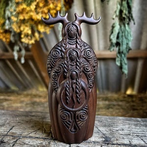 Puede incluir: Una talla de madera marrón oscuro de una figura con cornamenta, patrones intrincados y un diseño circular. La escultura tiene una superficie detallada y texturizada y se encuentra sobre una superficie de madera. La talla parece ser una representación de una deidad o figura mítica.