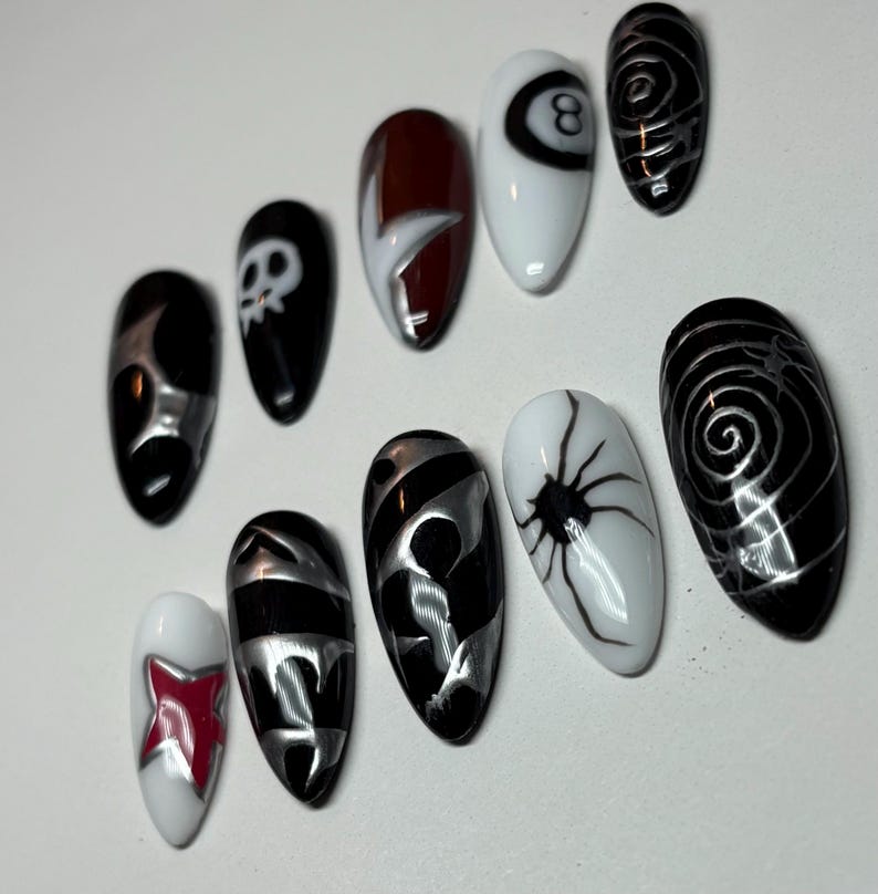K&ouml;nnte beinhalten: Ein Set aus zehn k&uuml;nstlichen N&auml;geln mit verschiedenen Designs. Die N&auml;gel weisen eine Mischung aus schwarzen, wei&szlig;en, silbernen und roten Farben auf. Designs umfassen einen Totenkopf, eine Spinne und abstrakte Muster. Die N&auml;gel sind oval geformt.