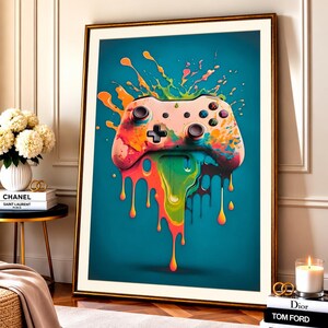 May include: A framed wall art print featuring a video game controller centred against a teal background. The controller is covered in vibrant, multi-coloured paint splatters and drips that extend downwards, creating a dynamic, fluid effect.
