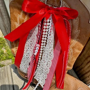 May include: A red ribbon boot charm with white lace, pearl beads, and red and white gingham ribbon. The charm is attached to a brown leather cowboy boot. The word "LOVE" is spelled out on the charm.