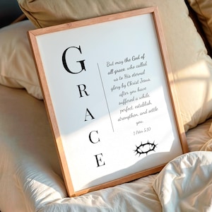 Puede incluir: Un cuadro enmarcado con la palabra "GRACE" en letras negras grandes. El cuadro incluye un versículo bíblico y una ilustración de una corona de espinas. El marco es de color marrón claro y el cuadro se muestra en una cama.