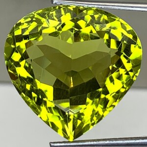 May include: A heart-shaped, faceted gemstone in a vivid, yellowish-green colour. Held by metal tweezers, the gem's intricate cut and clarity are highlighted. Light reflects off the facets, producing a sparkling effect. The shape and colour suggest a peridot or similar gemstone.