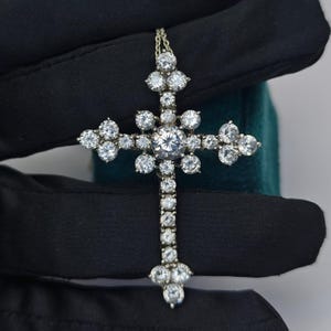 Puede incluir: Colgante de cruz de diamantes con una fina cadena de oro. La cruz presenta numerosos diamantes de talla redonda, con una piedra central más grande. El diseño es simétrico, con los diamantes dispuestos en un patrón floral.