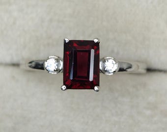 May include: A silver ring featuring a rectangular, deep red gemstone as the focal point, flanked by two smaller, round, clear gemstones. The ring's band is a simple, polished silver.
