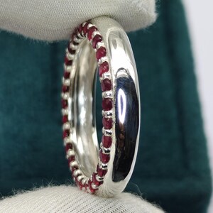 May include: A polished silver band ring featuring a continuous row of small, round, deep red rubies set along the inner edge. The metal surface is highly reflective, showing light highlights, while the gemstones create a vibrant red border.