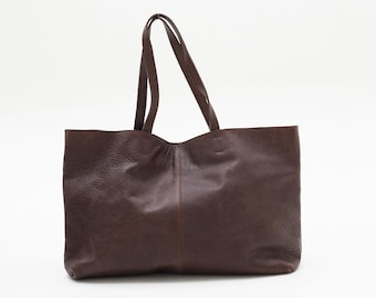 May include: A large, dark brown leather tote bag with two long handles. The bag has a rectangular shape with a slightly curved top edge and a textured surface. The handles are attached at the top and are made of the same leather material.