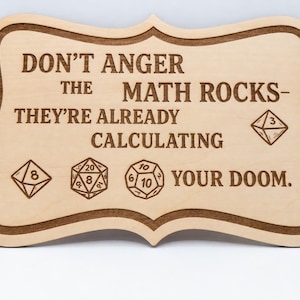 May include: A light-colored wooden sign with a decorative scalloped edge, featuring the engraved text: DON'T ANGER THE MATH ROCKS- THEY'RE ALREADY CALCULATING YOUR DOOM. The sign includes engraved illustrations of four different polyhedral dice.