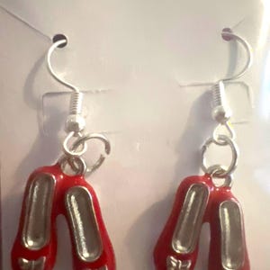 May include: A pair of red enamel earrings shaped like ballet slippers. Each slipper has a silver-colored interior and a small bow detail. The earrings hang from silver-colored fish hook ear wires.