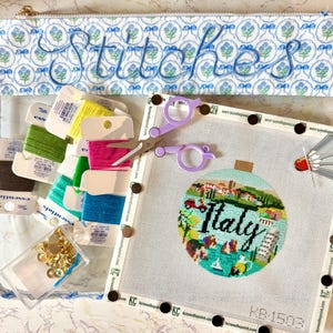 Puede incluir: Una bolsa transparente con cremallera con la palabra "Stitches" bordada en hilo azul. En el interior hay carretes de hilo colorido, cierres dorados y un lienzo de punto de aguja con un diseño de "Italia", junto con unas tijeras moradas y un alfiler decorativo.