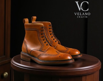 Puede incluir: Un par de botas de cuero color marrón claro con intrincados detalles de brogue y un diseño con cordones. Las botas tienen un estilo clásico con punta redondeada y suela resistente. Las botas se exhiben sobre una superficie de madera oscura. La marca "VELANO CRAFTS" es visible.