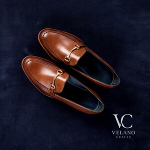 May include: A pair of brown leather loafers with gold-colored metal accents. The shoes are displayed on a dark blue textured surface. The brand logo "VC VELANO CRAFTS" is visible in the bottom right corner.