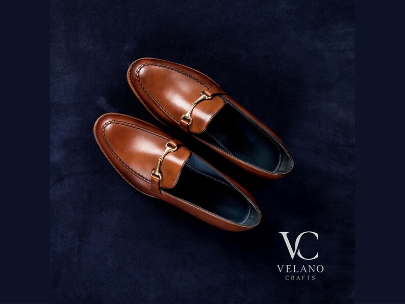 May include: A pair of brown leather loafers with gold-colored metal accents. The shoes are displayed on a dark blue textured surface. The brand logo "VC VELANO CRAFTS" is visible in the bottom right corner.