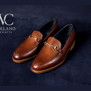 May include: A pair of brown leather loafers with gold-colored metal accents. The shoes have a classic design with a rounded toe and a slip-on style. The brand name "VELANO CRAFTS" is visible in the upper left corner.