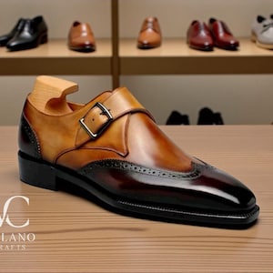 Könnte beinhalten: Zweifarbiges Lederschuh mit Schnallenverschluss. Der Schuh hat eine braune und schwarze Farbgebung mit dekorativen Perforationen. Das Logo "VC VELANO CRAFTS" ist auf der Holzoberfläche sichtbar.