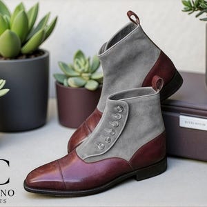 Puede incluir: Botines elegantes con parte superior de ante gris y detalles en cuero burdeos. Los botines tienen cierre de botones y una trabilla. Se exhiben con suculentas en macetas, mostrando una estética sofisticada.