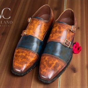 Pode incluir: Um par de sapatos de couro artesanais com um design único. Os sapatos apresentam uma combinação de cores laranja e azul marinho, com fecho de fivela. Os sapatos são exibidos numa superfície de madeira. A marca "VELANO CRAFTS" é visível.