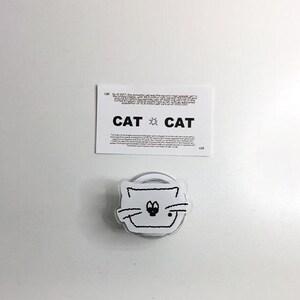 May include: A white cat-shaped phone grip with a black outline and a smiling face. Above it is a white card with the word "CAT" and text about cats. The background is white.