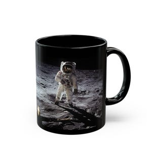 Peut inclure: Mug en céramique noire avec une image d'astronaute sur la lune. L'astronaute porte une combinaison spatiale blanche et se tient sur une surface lunaire grise. La tasse a une anse et un intérieur noirs.