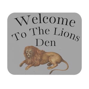 May include: A gray, rectangular sign with rounded corners. The text "Welcome To The Lions Den" is printed in black. A detailed illustration of a lion in shades of brown and tan is below the text.