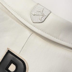 Puede incluir: Primer plano de una chaqueta de cuero blanca con un emblema cosido en el hombro. La chaqueta presenta un parche con una letra en blanco y negro. La costura y la textura del cuero son visibles.