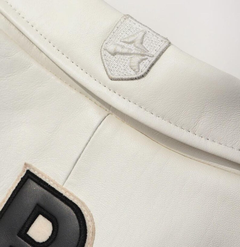 Puede incluir: Primer plano de una chaqueta de cuero blanca con un emblema cosido en el hombro. La chaqueta presenta un parche con una letra en blanco y negro. La costura y la textura del cuero son visibles.
