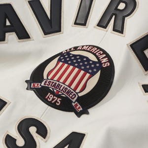 Puede incluir: Primer plano de una chaqueta blanca con letras y parches negros. La chaqueta presenta un parche con un dise&ntilde;o de bandera estadounidense y el texto "ALL AMERICANS ESTABLISHED 1975 N.Y."