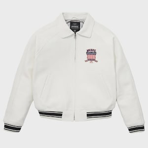 Puede incluir: Chaqueta blanca con cuello y cierre de cremallera. La chaqueta presenta un parche en el pecho con el texto "AVIREX" y un dise&ntilde;o en rojo, blanco y azul. Los pu&ntilde;os y el dobladillo tienen detalles a rayas blancas y negras.