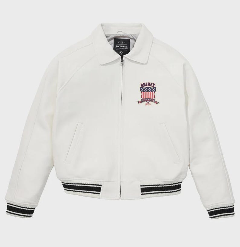Puede incluir: Chaqueta blanca con cuello y cierre de cremallera. La chaqueta presenta un parche en el pecho con el texto "AVIREX" y un dise&ntilde;o en rojo, blanco y azul. Los pu&ntilde;os y el dobladillo tienen detalles a rayas blancas y negras.