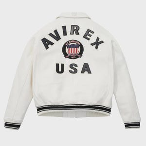 Puede incluir: Chaqueta de cuero blanca con pu&ntilde;os y dobladillo a rayas blancas y negras. La parte trasera presenta la palabra "AVIREX" sobre un emblema circular con las palabras "ALL AMERICANS ESTABLISHED 1975" y un dise&ntilde;o de bandera estadounidense. Debajo, la palabra "USA".