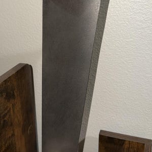 K&ouml;nnte beinhalten: Eine metallisch graue Titan-Statorschaufel mit einem glatten, sich verj&uuml;ngenden vertikalen K&ouml;rper und einer verst&auml;rkten Montagebasis an der Oberseite. Die Schaufel ist in einem rechteckigen Holzblock befestigt, auf dem TITANIUM STATOR BLADE eingraviert ist.