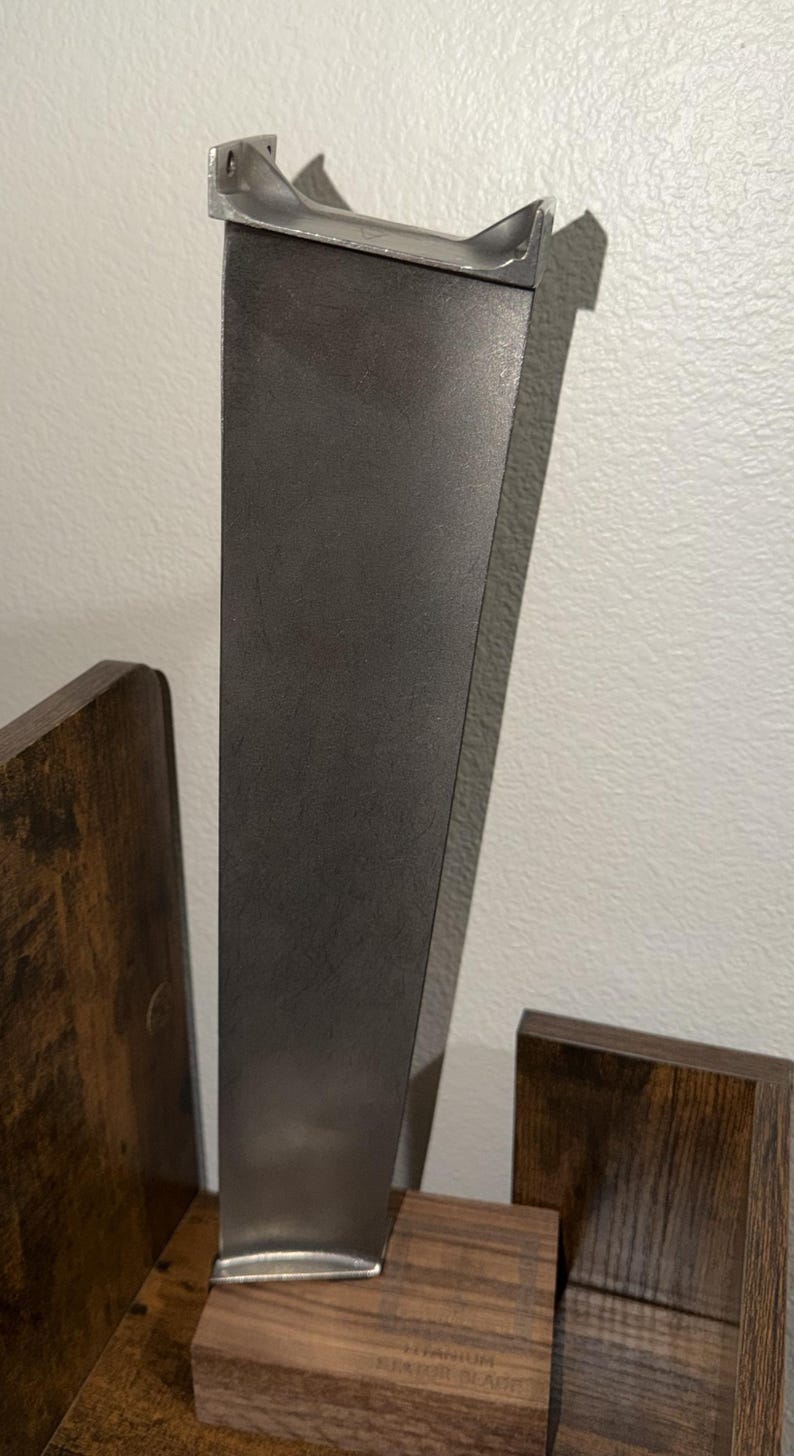 K&ouml;nnte beinhalten: Eine metallisch graue Titan-Statorschaufel mit einem glatten, sich verj&uuml;ngenden vertikalen K&ouml;rper und einer verst&auml;rkten Montagebasis an der Oberseite. Die Schaufel ist in einem rechteckigen Holzblock befestigt, auf dem TITANIUM STATOR BLADE eingraviert ist.
