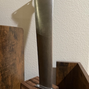 K&ouml;nnte beinhalten: Eine metallische Turbinenschaufel mit glattem, sich verj&uuml;ngendem vertikalem Design, die mit zwei kleinen Metallschrauben auf einem rechteckigen Holzsockel befestigt ist. Die Schaufel hat eine matte silberne Oberfl&auml;che und eine schr&auml;ge Oberkante.