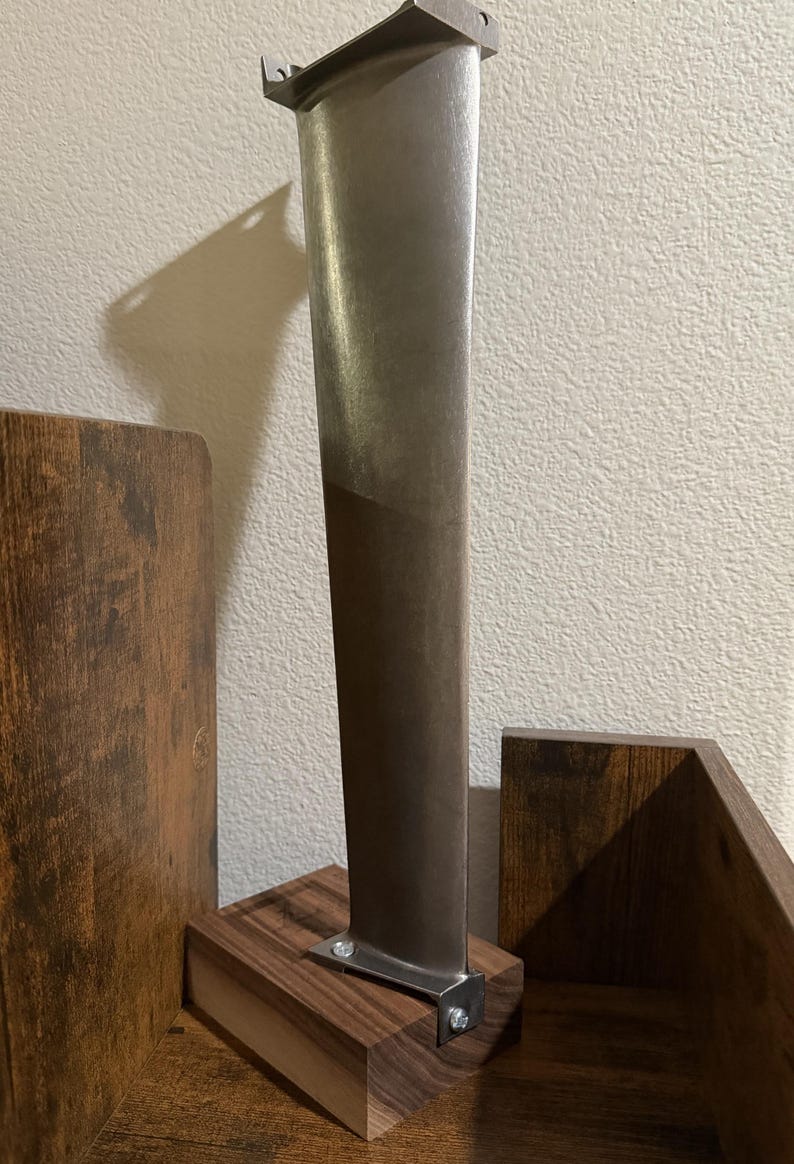 K&ouml;nnte beinhalten: Eine metallische Turbinenschaufel mit glattem, sich verj&uuml;ngendem vertikalem Design, die mit zwei kleinen Metallschrauben auf einem rechteckigen Holzsockel befestigt ist. Die Schaufel hat eine matte silberne Oberfl&auml;che und eine schr&auml;ge Oberkante.