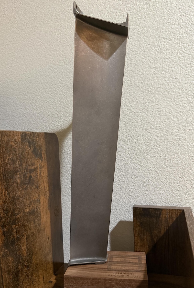 K&ouml;nnte beinhalten: Eine hohe, metallische, graue Turbinen-Statorschaufel mit einem glatten, sich verj&uuml;ngenden vertikalen Design. Die Schaufel ist auf einem rechteckigen Holzsockel montiert, der die Gravur Titanium Stator Blade tr&auml;gt und industriell wirkt.