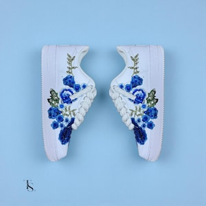 Pode incluir: Par de tênis brancos com intrincado bordado floral azul. O design apresenta flores azuis detalhadas, folhas verdes e detalhes dourados. Os cadarços são brancos e o fundo é azul claro.