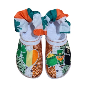 Op de afbeelding: Witte klompen versierd met oranje en gouden strass, klavers en een hart in de kleuren van de Ierse vlag. Eén klomp heeft een kabouter met een pot goud, de andere heeft een Guinness-logo. De klompen zijn afgewerkt met groene, witte en oranje stof.