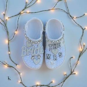 Op de afbeelding: Een paar witte Crocs versierd met parels en strass. Eén schoen zegt "THE MRS" en de andere "THE BRIDE" in parelletters. De schoenen hebben hart- en kroondetails.