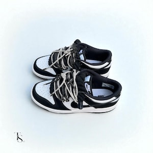 May include: A pair of black and white Nike Dunk Low sneakers with thick, rope-like laces. The shoes have a white base with black overlays and a white midsole. The Nike logo is visible on the tongue and side of the shoe.