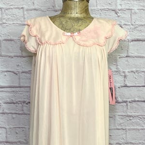 May include: A vintage, pale pink nightgown with a scalloped neckline and hem. The nightgown features a delicate pink bow and floral embellishment on the bodice. The short sleeves and hem are trimmed with a darker pink trim. The nightgown is displayed on a mannequin.