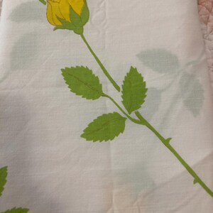 Op de afbeelding: Witte stof met een herhaald patroon van gele rozen, groene bladeren en stelen. De rozen staan in volle bloei en de bladeren hebben een gedetailleerd, gestructureerd uiterlijk. De stof is waarschijnlijk een laken of ander huishoudtextiel.