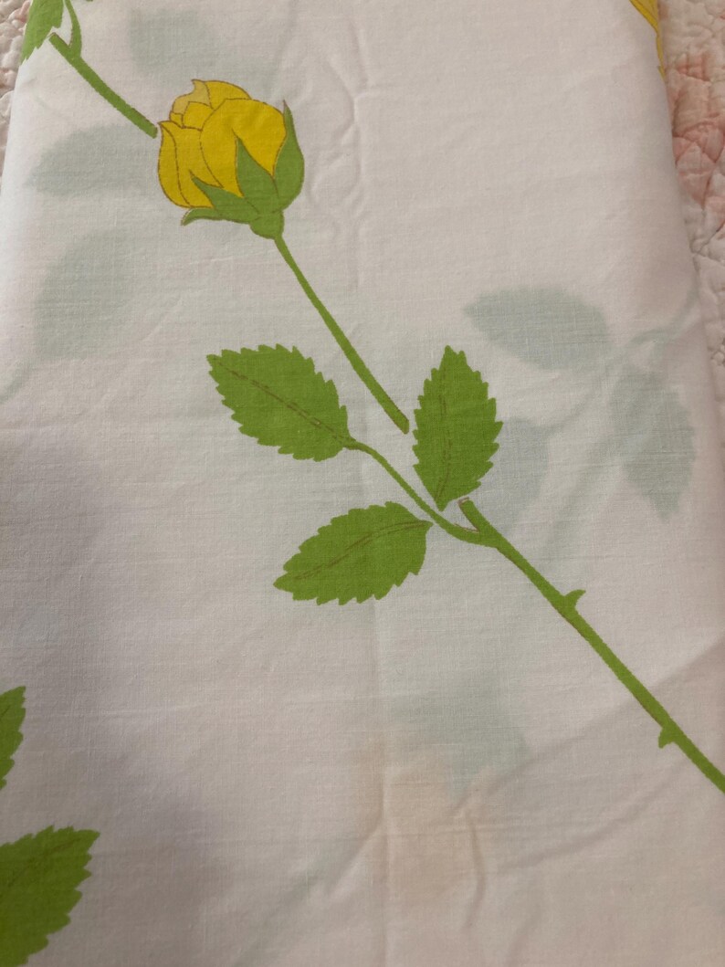 Op de afbeelding: Witte stof met een herhaald patroon van gele rozen, groene bladeren en stelen. De rozen staan in volle bloei en de bladeren hebben een gedetailleerd, gestructureerd uiterlijk. De stof is waarschijnlijk een laken of ander huishoudtextiel.