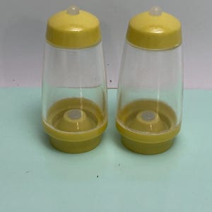 May include: Two clear plastic salt and pepper shakers with yellow lids and bases. The shakers have a classic design, with a rounded base and a slightly tapered body. The lids have a small, clear, rounded top.