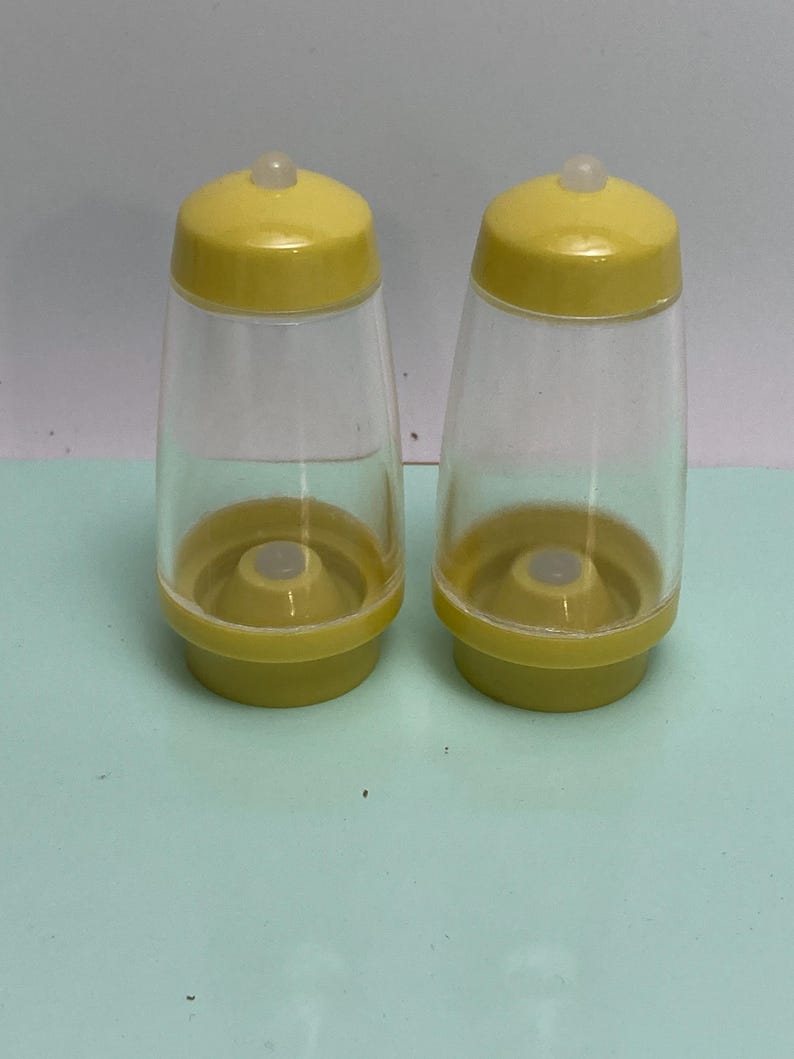 May include: Two clear plastic salt and pepper shakers with yellow lids and bases. The shakers have a classic design, with a rounded base and a slightly tapered body. The lids have a small, clear, rounded top.