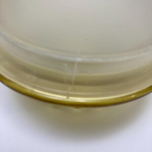 May include: A clear, round, plastic container lid with a slightly frosted appearance. The lid has a yellow-tinted rim and a slightly domed center. The container is likely for food storage.
