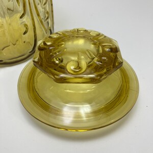 May include: A vintage yellow glass jar lid and base. The lid is octagonal with a floral design, and the base is round. The glass has a slight amber tint. The jar is likely for kitchen storage.