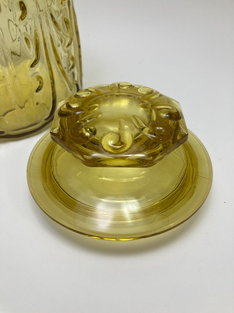 May include: A vintage yellow glass jar lid and base. The lid is octagonal with a floral design, and the base is round. The glass has a slight amber tint. The jar is likely for kitchen storage.