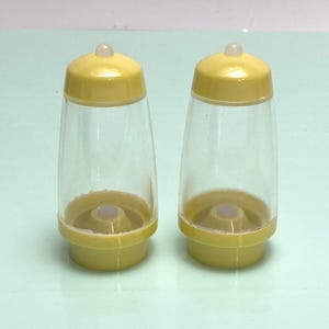 May include: A pair of yellow and clear plastic salt and pepper shakers. Each shaker has a yellow base and lid, with a clear, hourglass-shaped body. The lids have a small, rounded top. The shakers are designed for tabletop use.