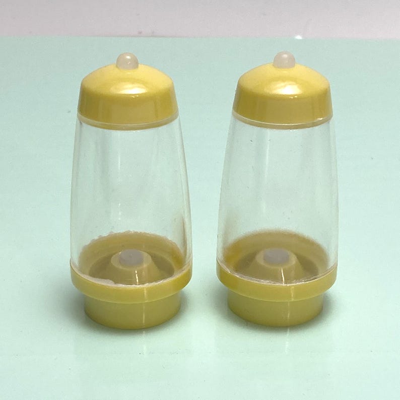 May include: A pair of yellow and clear plastic salt and pepper shakers. Each shaker has a yellow base and lid, with a clear, hourglass-shaped body. The lids have a small, rounded top. The shakers are designed for tabletop use.