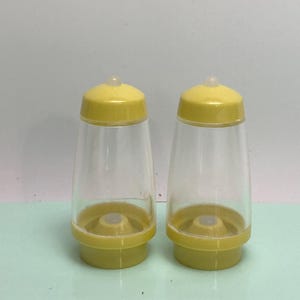 May include: Two clear plastic salt and pepper shakers with yellow lids and bases. The shakers have a conical shape and a small, clear plastic top on the lid. The bases are a solid yellow color.