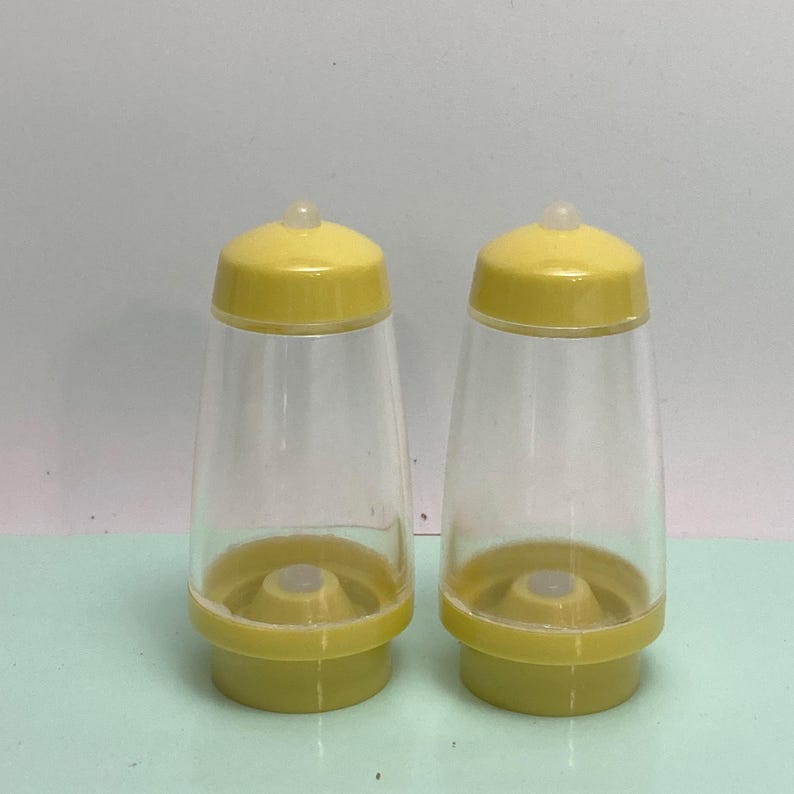 May include: Two clear plastic salt and pepper shakers with yellow lids and bases. The shakers have a conical shape and a small, clear plastic top on the lid. The bases are a solid yellow color.